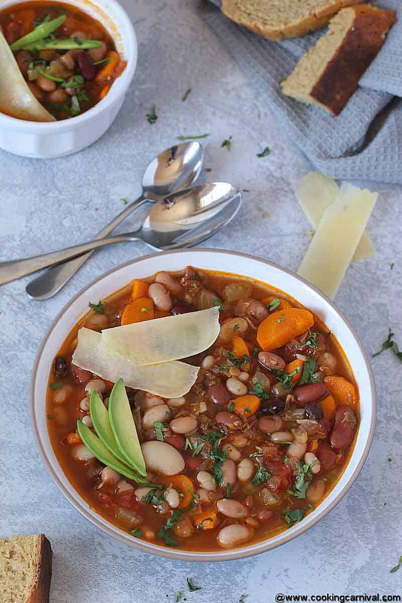 Vegetarian 15 beans soup in white bowl with Parmesan cheese, cilantro and avocado, 2 spoons and some crusty bread on the side